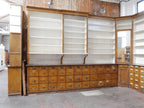 Schräg aufgenommene Ansicht des linken Bereichs der historischen Apothekeneinrichtung mit offenen Wandregalen, originalen Unterschränken und zahlreichen Schubladen aus massiver Kiefer.