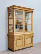 Schräg aufgenommene Frontansicht der historischen Bibliotheksvitrine aus Weichholz. Gut erkennbar sind die originalen Glasscheiben, die massiven Füllungstüren im Unterteil sowie die elegante Bauweise des ehemaligen Ladenschranks.