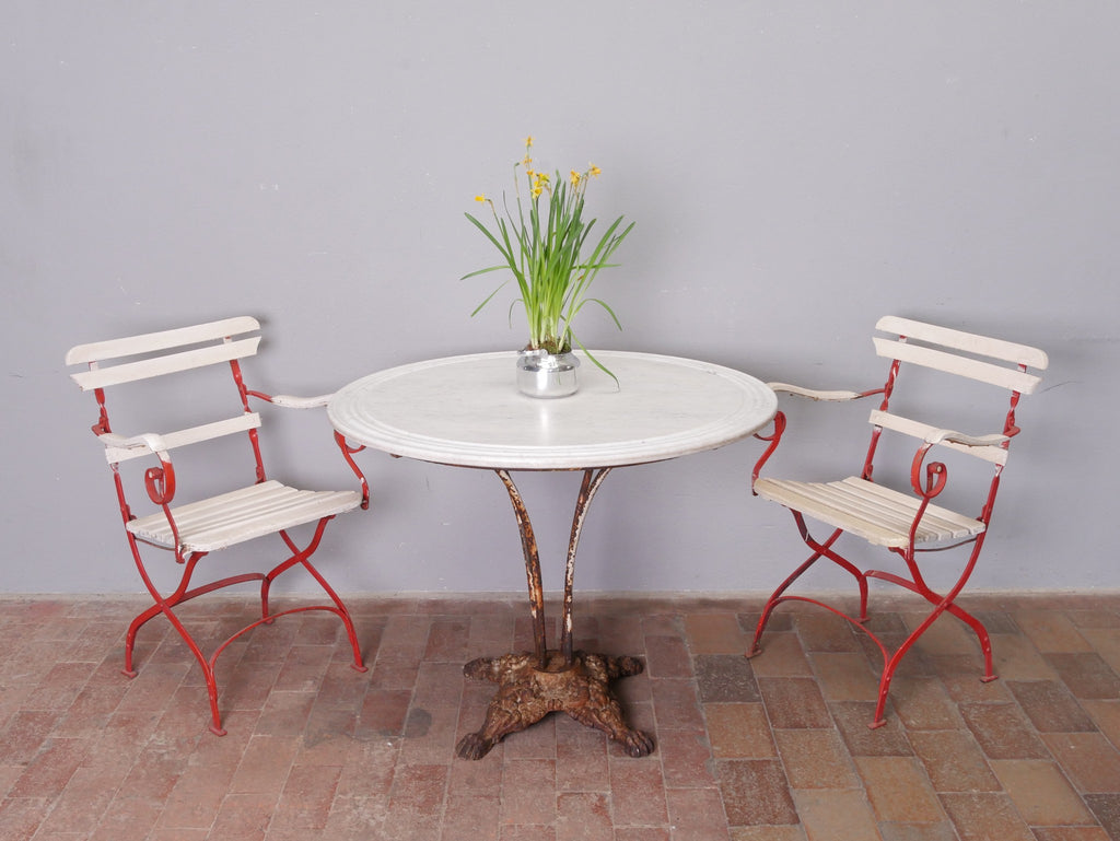 Historischer Gartentisch mit originaler Marmorplatte und kunstvoll gearbeitetem Gusseisengestell, dekoriert mit einer Vase aus Metall und blühenden Narzissen. Ergänzt wird das Ensemble durch zwei originale Gartenstühle mit weißen Holzlatten und roten Eisengestellen. Die Aufnahme vermittelt die elegante Wirkung des antiken Gartenmöbels im dekorierten Zustand.