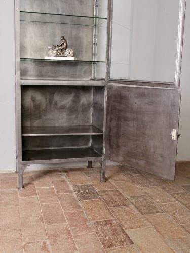 Schräg aufgenommener Arzt- und Büroschrank mit geöffneter Glastür. Die Kombination aus verglastem Oberteil und geschlossenem Unterschrank sowie die restaurierte Metalloberfläche zeigen den Schrank als funktionales und dekoratives Originalmöbel aus der Zeit um 1910–1920.
