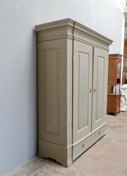 Schräg von der rechten Seite fotografierter Empire Kleiderschrank mit markanter Kranzleiste, Seitenfüllungen und moderner grauer Neufassung.