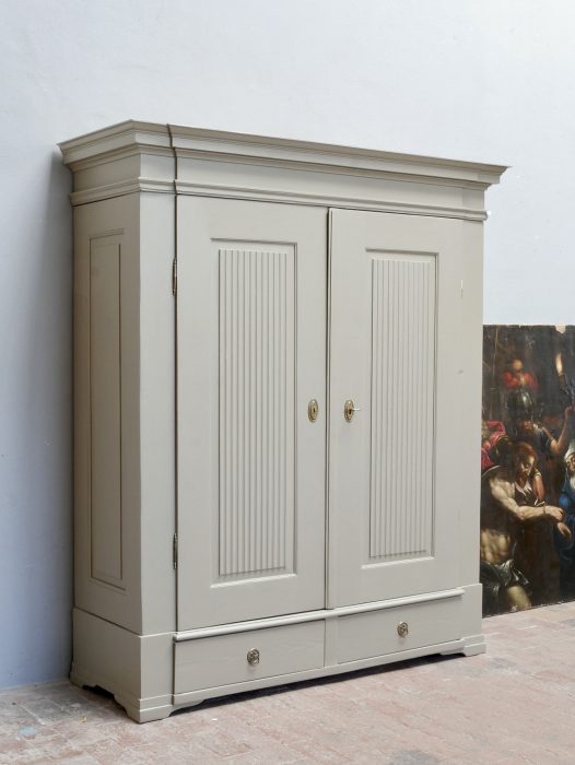 Großer originaler Empire Kleiderschrank bzw. Wäscheschrank um 1800 in der Hauptansicht. Der beeindruckende zweitürige Schrank aus Weichholz besitzt eine moderne graue Neufassung, markante Kannelierungen in den Türen, große Seitenfüllungen, außenliegende Scharniere und zwei geräumige Schubladen im Sockelbereich.