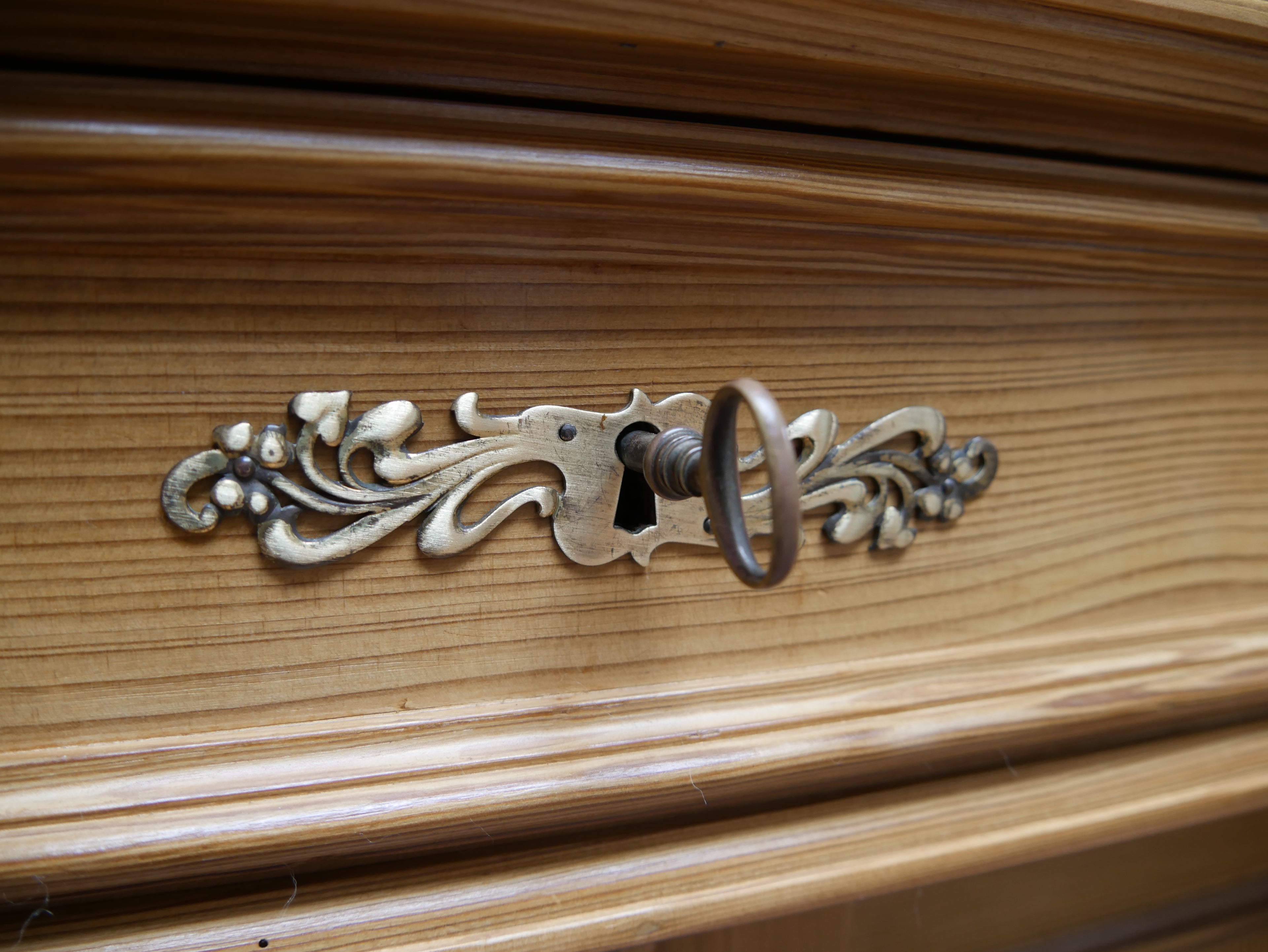 Detailaufnahme des kunstvoll gestalteten Originalbeschlags am restaurierten Gründerzeit-Vertiko aus Weichholz, um 1870. Der historische Metallbeschlag ist floral und geschwungen ornamentiert und umfasst das zentrale Schlüsselloch mit eingesetztem Originalschlüssel. Im Hintergrund ist die fein gemaserte und gewachste Weichholzoberfläche der Möbelplatte mit profilierten Holzleisten zu sehen. 