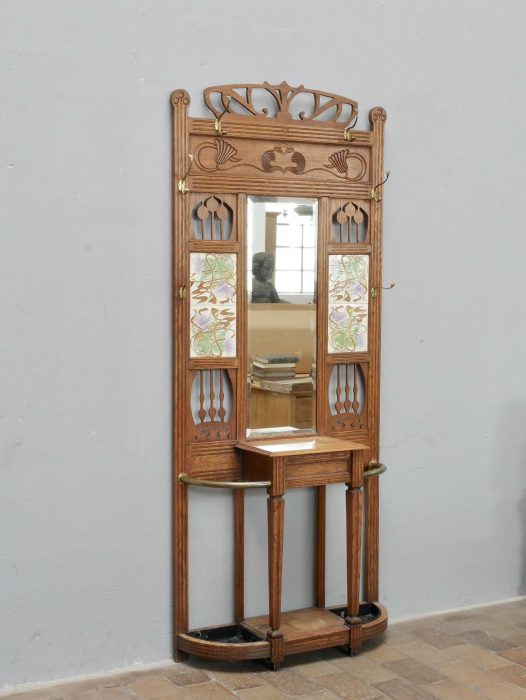 Gesamtansicht der originalen Jugendstil-Garderobe aus leicht linker Perspektive. Gut erkennbar sind die elegante Gesamtkonstruktion, die dekorative Krone, der Spiegel, die Keramikfliesen sowie die beiden Schirmständer im Sockelbereich.
