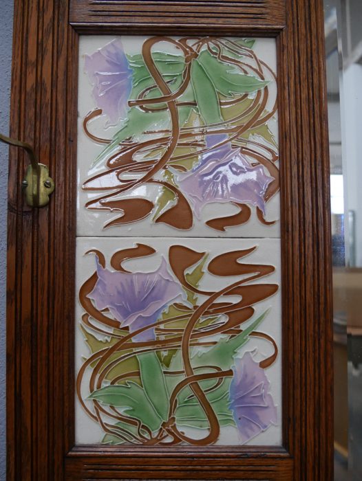 Detailansicht einer originalen Jugendstil-Keramikfliese mit floralem Dekor in Grün- und Lilatönen. Die kunstvolle Fliese gehört zur originalen Ausstattung der Garderobe und ist hervorragend erhalten.