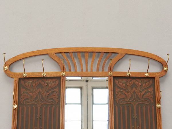Detailaufnahme des oberen Bereichs mit geschwungenem Jugendstil-Aufsatz, großem Facettenspiegel sowie den seitlichen Garderobenhaken. Die elegante Linienführung prägt das repräsentative Erscheinungsbild der Garderobe.
