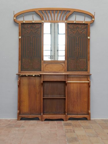 Frontalansicht der kompletten Jugendstil-Garderobe mit großem Facettenspiegel, geprägten Lederfüllungen, seitlichen Garderobenhaken, Ablageflächen und dekorativem Aufsatz. Die harmonischen Proportionen und die aufwendige Gestaltung spiegeln den typischen Jugendstil um 1900 wider.
