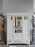Frontansicht des farbig gefassten Jugendstil-Kleiderschranks mit geschlossenen Türen. Gut sichtbar sind der originale Facettenspiegel, die kleinen Spiegel in den Außentüren, die eleganten Kassettenfüllungen sowie die beiden großen Sockelschubladen.
