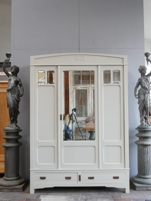 Frontansicht des farbig gefassten Jugendstil-Kleiderschranks mit geschlossenen Türen. Gut sichtbar sind der originale Facettenspiegel, die kleinen Spiegel in den Außentüren, die eleganten Kassettenfüllungen sowie die beiden großen Sockelschubladen.
