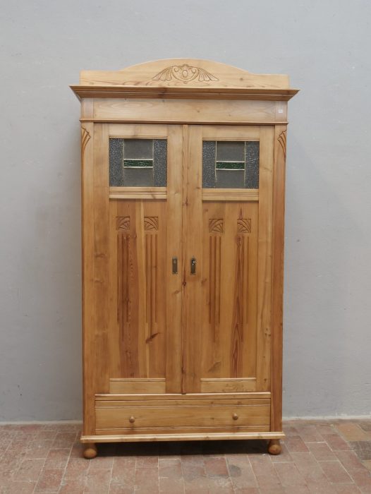 Frontansicht des restaurierten Jugendstil-Kleiderschranks mit originalen Bleiglasfenstern, geschnitzten Türfüllungen und dekorativem Giebelaufsatz. Die harmonischen Proportionen und die gewachste Weichholzoberfläche unterstreichen den eleganten Charakter des historischen Schranks.
