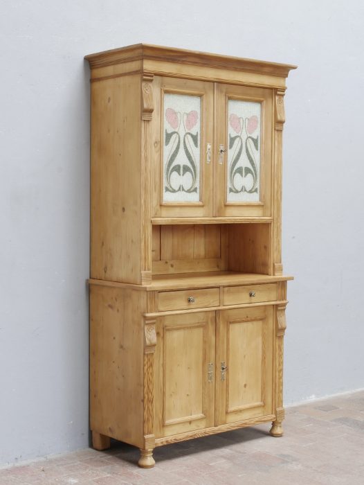 Schräge Vorderansicht des restaurierten Jugendstil-Küchenbuffetts aus Weichholz. Die harmonischen Proportionen, die zeittypischen Verzierungen und die original bemalten Glasscheiben im Oberteil sind gut erkennbar.