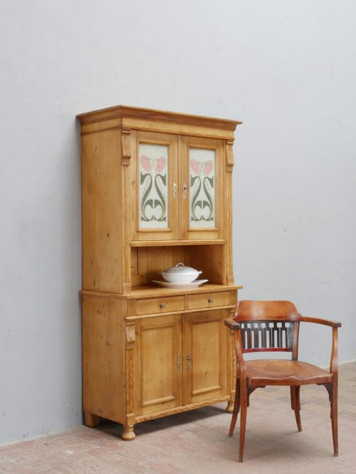 Hauptansicht eines restaurierten Jugendstil-Küchenbuffetts beziehungsweise einer Anrichte aus Weichholz, original um 1900. Das zweiteilige Möbel mit offenen Ablageflächen, Unterschrank, Schubladen und kunstvoll bemalten Original-Glasscheiben wird mit einem Stuhl als Größenvergleich präsentiert.