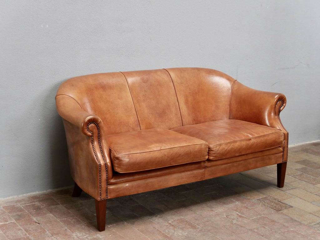 Schräg von links aufgenommenes Ledersofa aus cognacfarbenem Schafsleder. Die elegante Form mit gerundeter Rückenlehne, geschwungenen Armlehnen und der authentischen Lederpatina kommt besonders gut zur Geltung.
