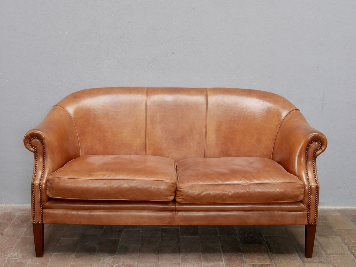 Frontansicht des Vintage-Ledersofas aus Schafsleder mit zwei großzügigen Sitzpolstern, elegant geschwungener Rückenlehne und warmem Cognac-Farbton. Die gleichmäßige Patina unterstreicht den authentischen Originalzustand.
