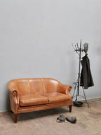 Exklusives Zweisitzer-Ledersofa aus den Niederlanden um 1970 aus hochwertigem Schafsleder. Das Sofa mit geschwungenen Armlehnen, wunderschöner Patina und warmem Cognac-Farbton ist als stilvolles Lounge- und Loftmöbel in einer Gesamtansicht mit Garderobenständer dekoriert.
