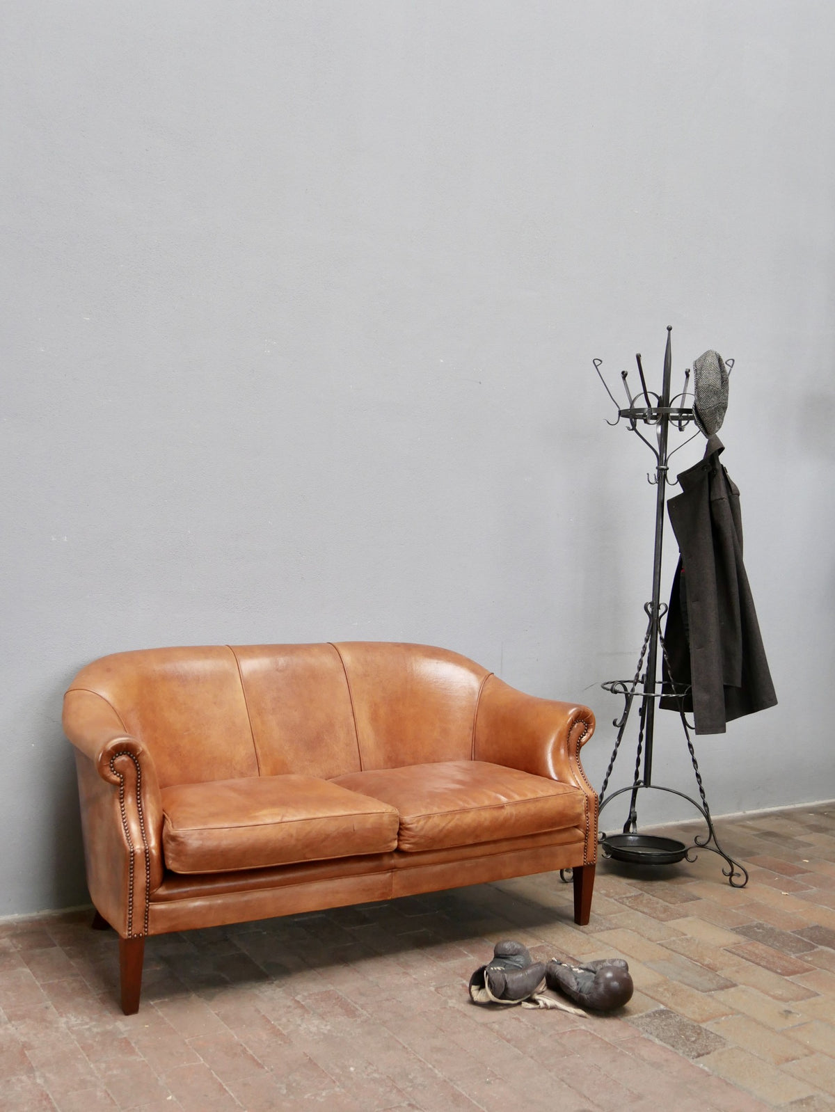 Exklusives Zweisitzer-Ledersofa aus den Niederlanden um 1970 aus hochwertigem Schafsleder. Das Sofa mit geschwungenen Armlehnen, wunderschöner Patina und warmem Cognac-Farbton ist als stilvolles Lounge- und Loftmöbel in einer Gesamtansicht mit Garderobenständer dekoriert.
