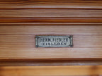 Nahaufnahme des originalen Herstellerschildes der Firma Hermann Fiedler aus Eisleben. Das erhaltene Metallschild dokumentiert die Herkunft und Authentizität der historischen Lehrmittelvitrine.