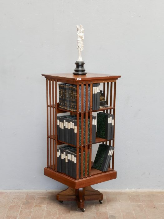 Schräge Frontansicht des drehbaren Jugendstil-Bücherkarussells aus massivem Nussbaum. Gut sichtbar sind die offenen Bücherfächer, die elegante Konstruktion mit senkrechten Streben sowie der massive drehbare Mittelfuß.
