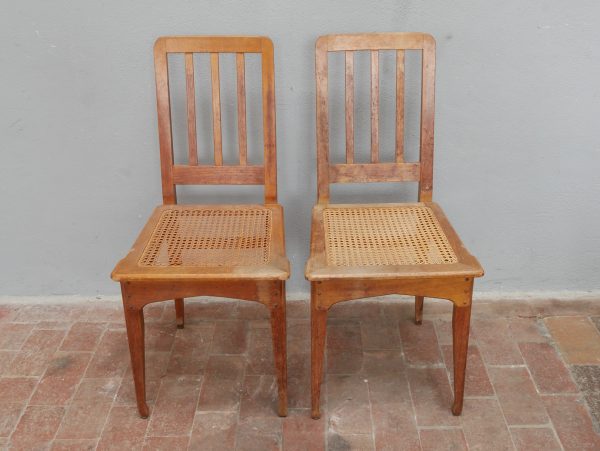 Frontalansicht zweier originaler Jugendstil-Stühle aus dem sechsteiligen Satz. Die Aufnahme zeigt die identische Ausführung, die senkrechten Rückenleisten sowie die originalen Sitzflächen mit Rohrgeflecht.
