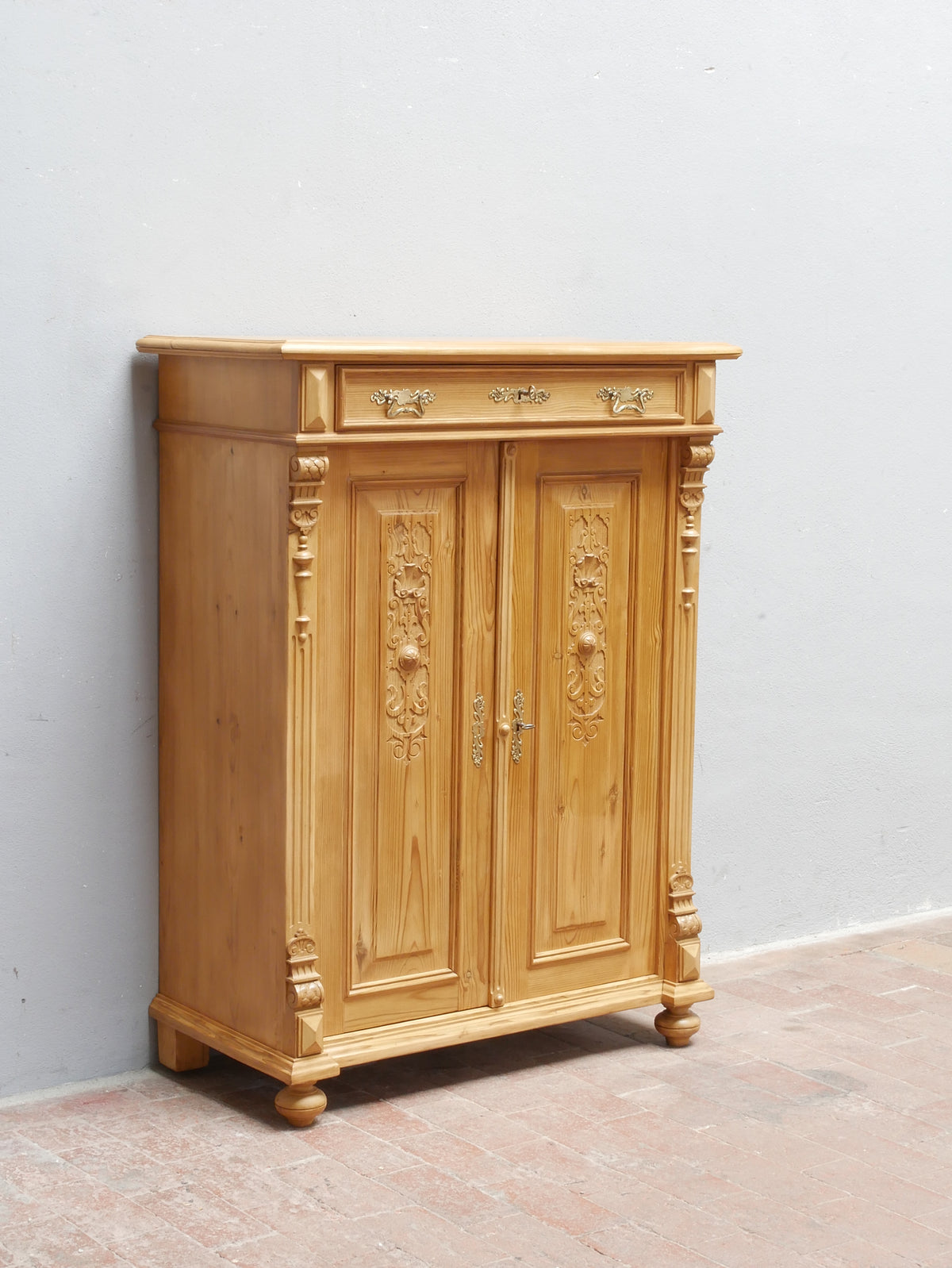 Sehr seltenes Gründerzeit-Vertiko aus Weichholz, um 1870, in restauriertem Zustand. Das aufwendig gestaltete historische Vertiko zeigt eine symmetrische Front mit zwei Türen, geschnitzten zeittypischen Gründerzeit-Ornamenten, dekorativen Originalbeschlägen und profiliertem oberen Abschluss. Die Oberfläche ist gewachst und bringt die warme Holzstruktur deutlich zur Geltung. Das Möbel steht vor einer hellen Wand auf einem gefliesten Boden und ist vollständig von vorne zu sehen.