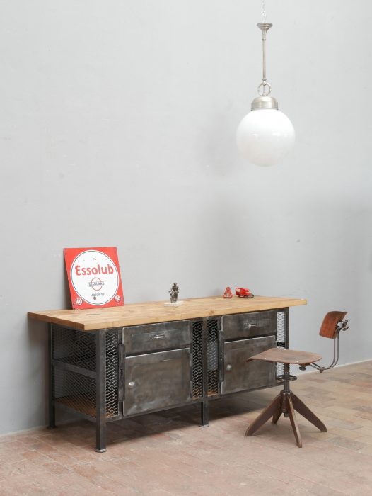 Hauptansicht einer restaurierten historischen Werkbank im Industriedesign mit massiver originaler Holzplatte und Metallkorpus. Die Werkbank eignet sich als Kücheninsel, Werktisch, Ladeneinrichtung oder Loft-Möbel und wird zusammen mit einem Industriehocker als Größenvergleich gezeigt.