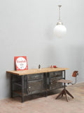 Hauptansicht einer restaurierten historischen Werkbank im Industriedesign mit massiver originaler Holzplatte und Metallkorpus. Die Werkbank eignet sich als Kücheninsel, Werktisch, Ladeneinrichtung oder Loft-Möbel und wird zusammen mit einem Industriehocker als Größenvergleich gezeigt.