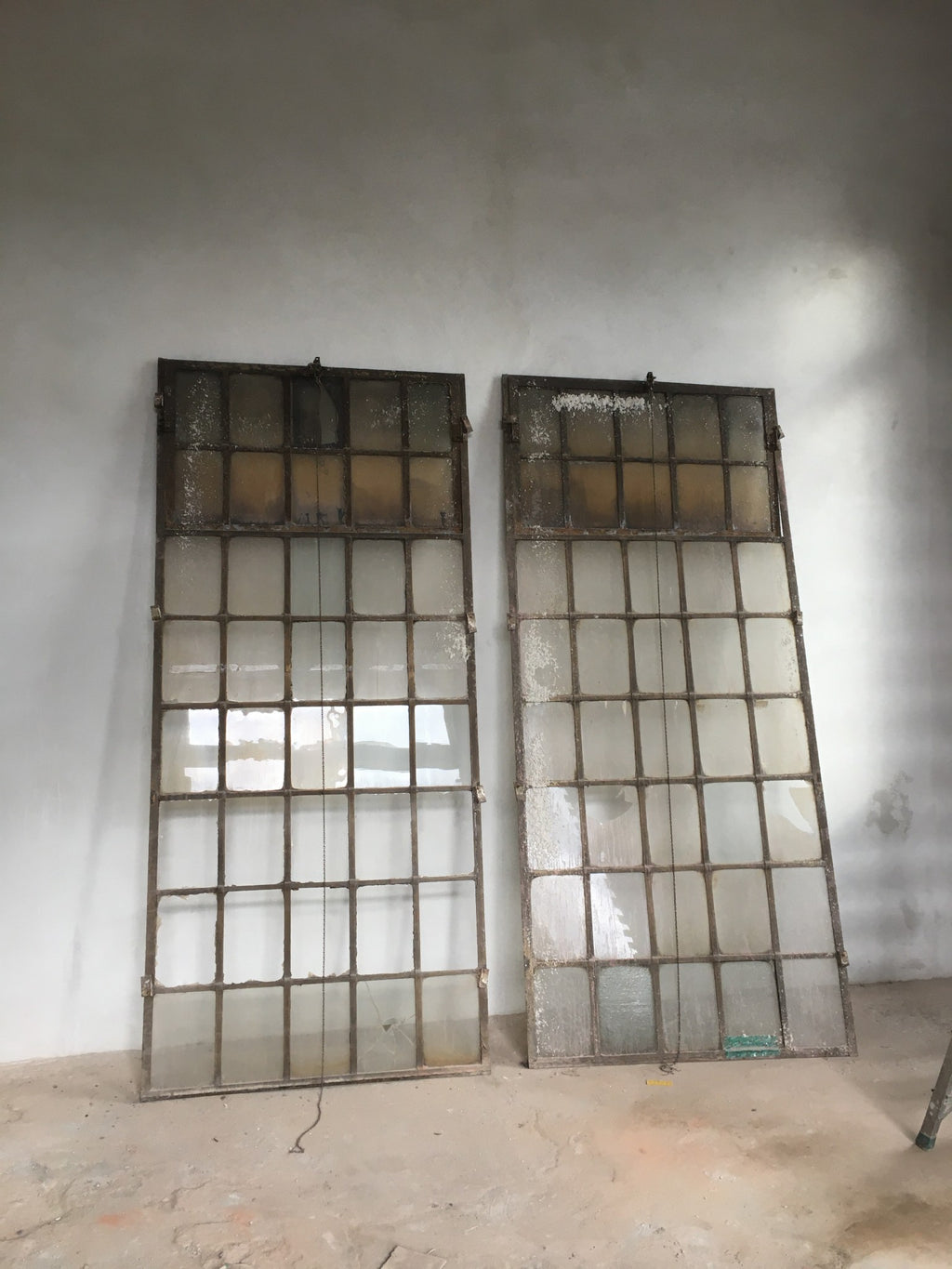 Zwei originale Fabrikfenster aus Schmiedeeisen um 1900 in der Gesamtansicht. Gut erkennbar sind die filigranen Sprossen, die großzügigen Glasflächen sowie die oberen Oberlichter, die über eine Kettenmechanik geöffnet werden können. Historische Industriefenster für Sanierung, Denkmalpflege oder exklusive Bauprojekte mit authentischem Fabrikcharakter.