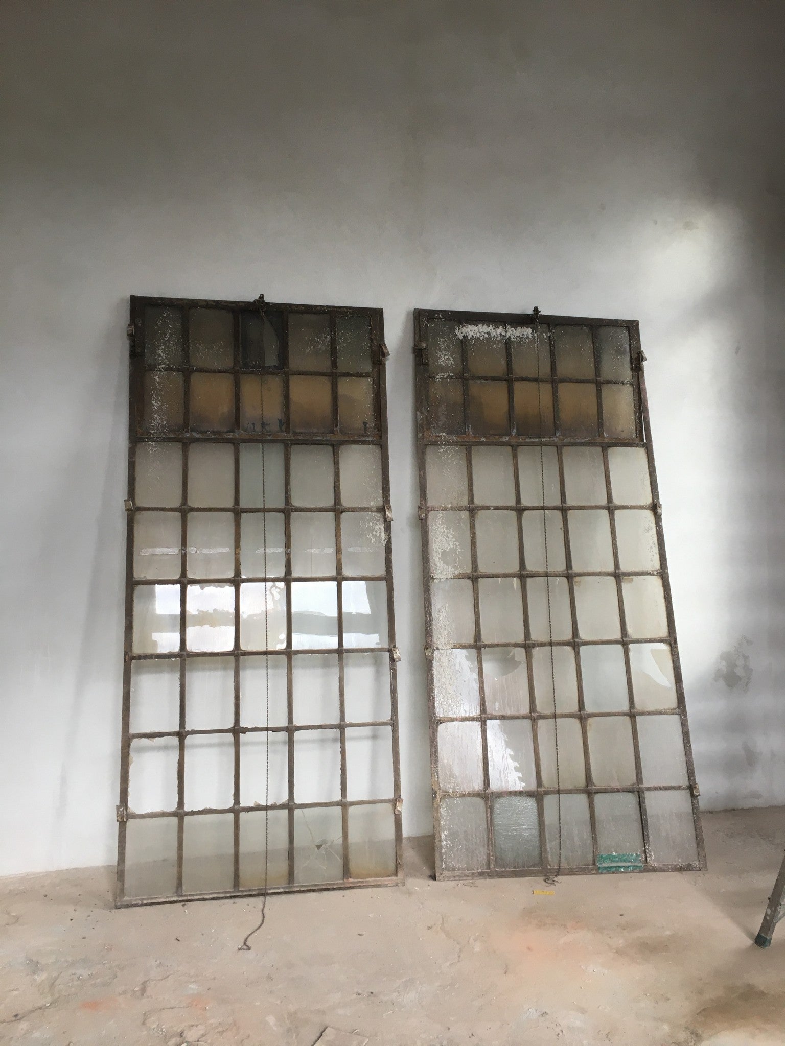 Zwei originale Fabrikfenster aus Schmiedeeisen um 1900 in der Gesamtansicht. Gut erkennbar sind die filigranen Sprossen, die großzügigen Glasflächen sowie die oberen Oberlichter, die über eine Kettenmechanik geöffnet werden können. Historische Industriefenster für Sanierung, Denkmalpflege oder exklusive Bauprojekte mit authentischem Fabrikcharakter.