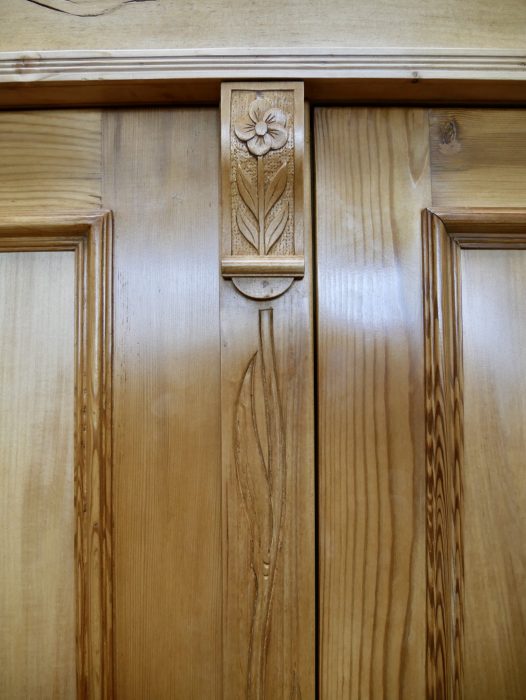 Detailaufnahme der mittleren Türpartie des restaurierten XXXL Jugendstil-Kleiderschranks aus gewachster Kiefer um 1900. Zu sehen ist das fein von Hand geschnitzte florale Jugendstil-Ornament mit stilisierter Blüte und langem Stängel auf dem mittleren Türsteg. Die hochwertige Schnitzarbeit, die natürliche Holzmaserung und die originalgetreue Restaurierung unterstreichen die außergewöhnliche handwerkliche Qualität dieses historischen Kleider- und Wäscheschranks.