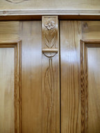 Detailaufnahme der mittleren Türpartie des restaurierten XXXL Jugendstil-Kleiderschranks aus gewachster Kiefer um 1900. Zu sehen ist das fein von Hand geschnitzte florale Jugendstil-Ornament mit stilisierter Blüte und langem Stängel auf dem mittleren Türsteg. Die hochwertige Schnitzarbeit, die natürliche Holzmaserung und die originalgetreue Restaurierung unterstreichen die außergewöhnliche handwerkliche Qualität dieses historischen Kleider- und Wäscheschranks.