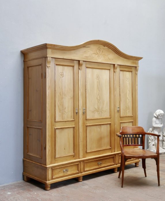 Hauptansicht eines beeindruckenden XXXL Jugendstil-Kleider- und Wäscheschranks aus gewachster Kiefer um 1900. Der restaurierte dreitürige Schrank besitzt einen geschweiften Giebel, geschnitzte Türfüllungen, drei große Schubladen im Sockelbereich und originale Beschläge. Ein Armlehnstuhl dient als Größenvergleich.